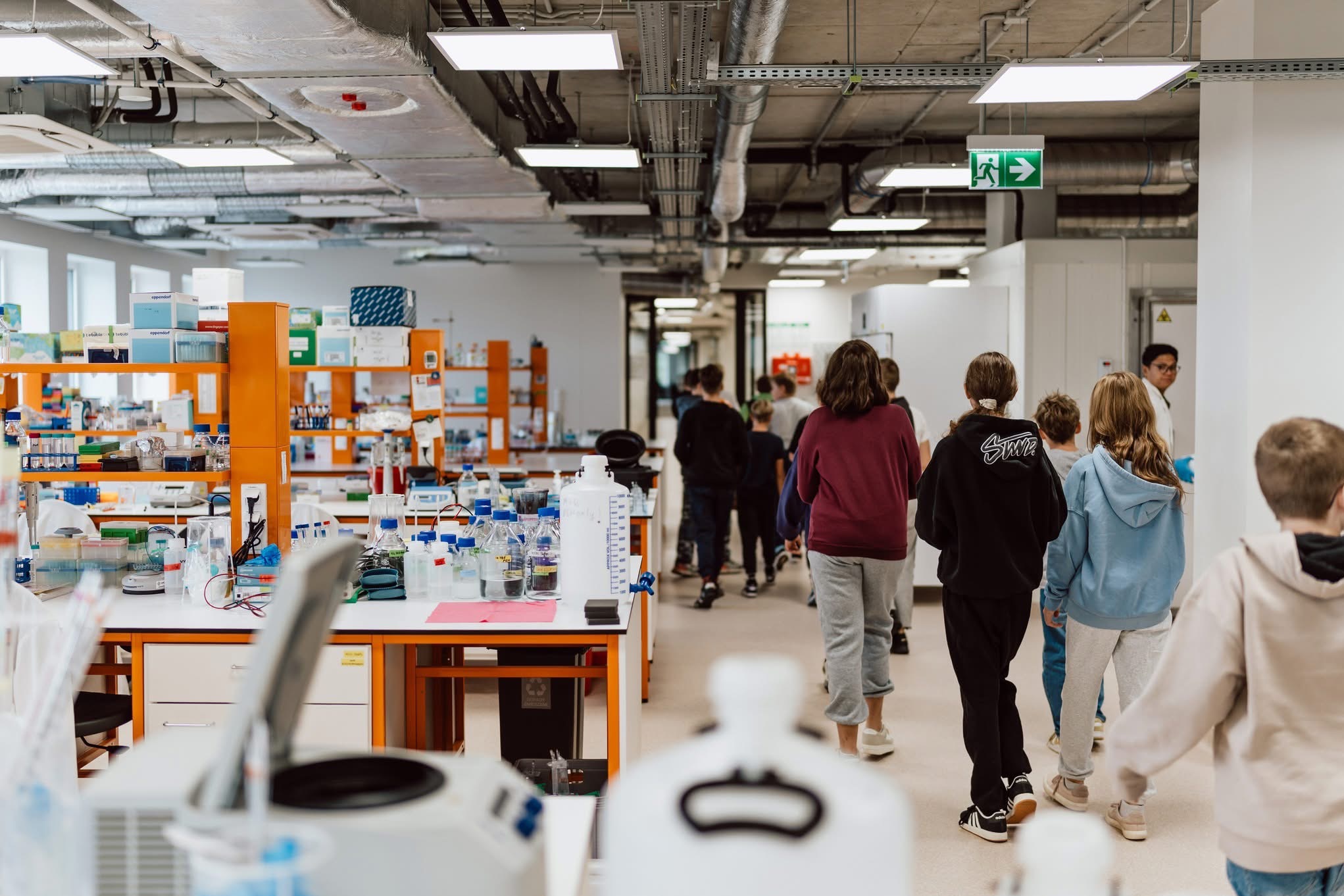 Grupa uczniów w laboratorium naukowym, widok z tyłu, z pomarańczowymi stołami i sprzętem laboratoryjnym. Oświetlenie sufitowe i znak ewakuacyjny.