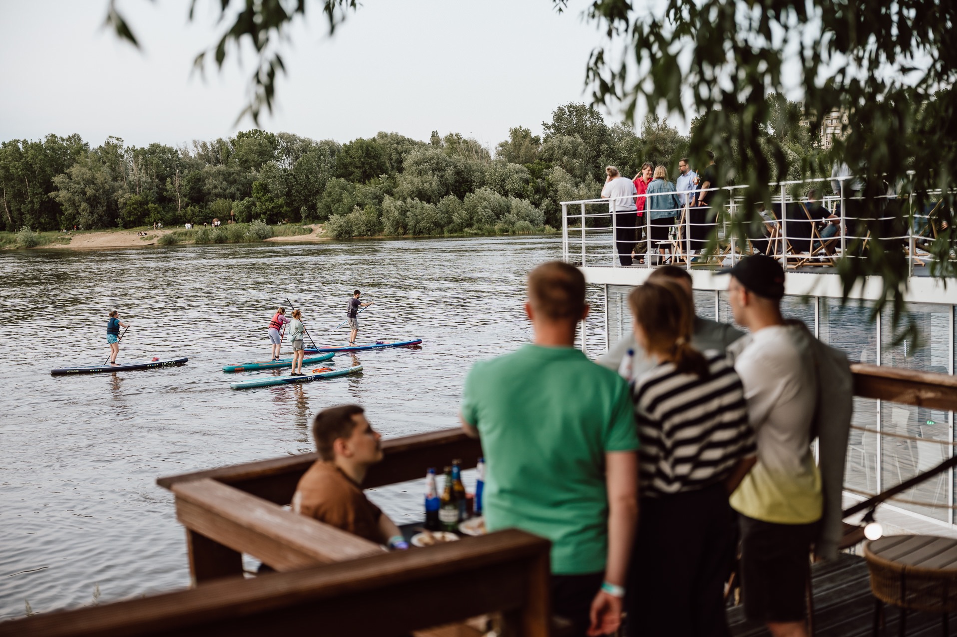Event na wodzie: grupa osób na deskach SUP, obserwatorzy z platformy. Widok z drewnianego tarasu, zieleń w tle, pochmurne niebo. Atmosfera relaksu i zabawy.