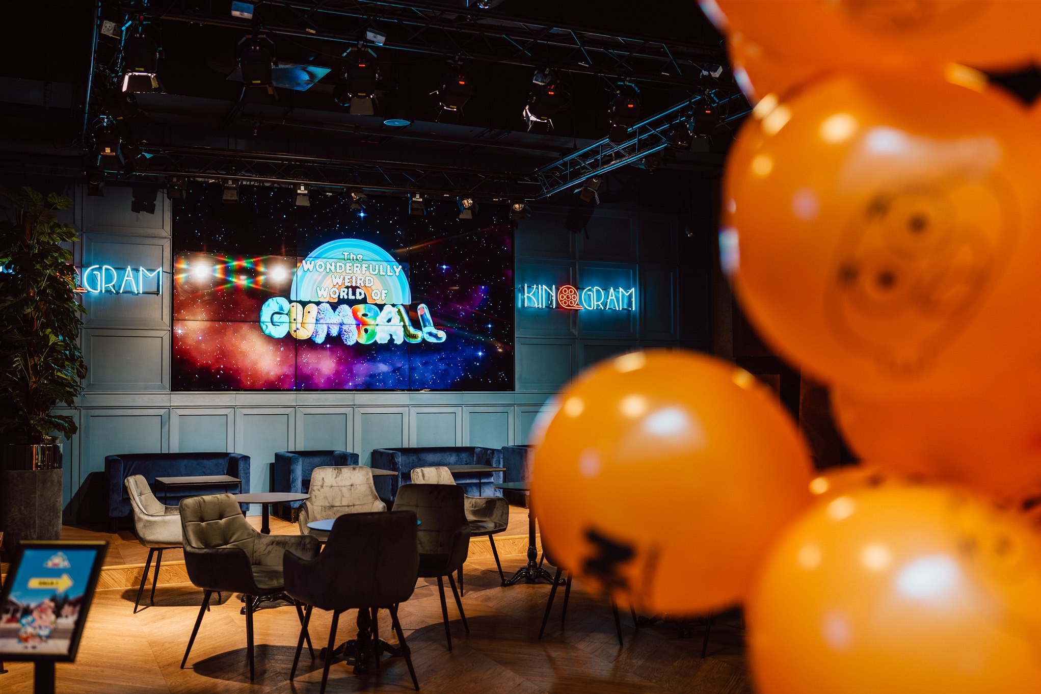 Sala eventowa z neonami 'GRAM' i ekranem 'Gumball', dekorowana pomarańczowymi balonami. Ustawione stoliki i sofy. Scena z oświetleniem. Unikalny wystrój.
