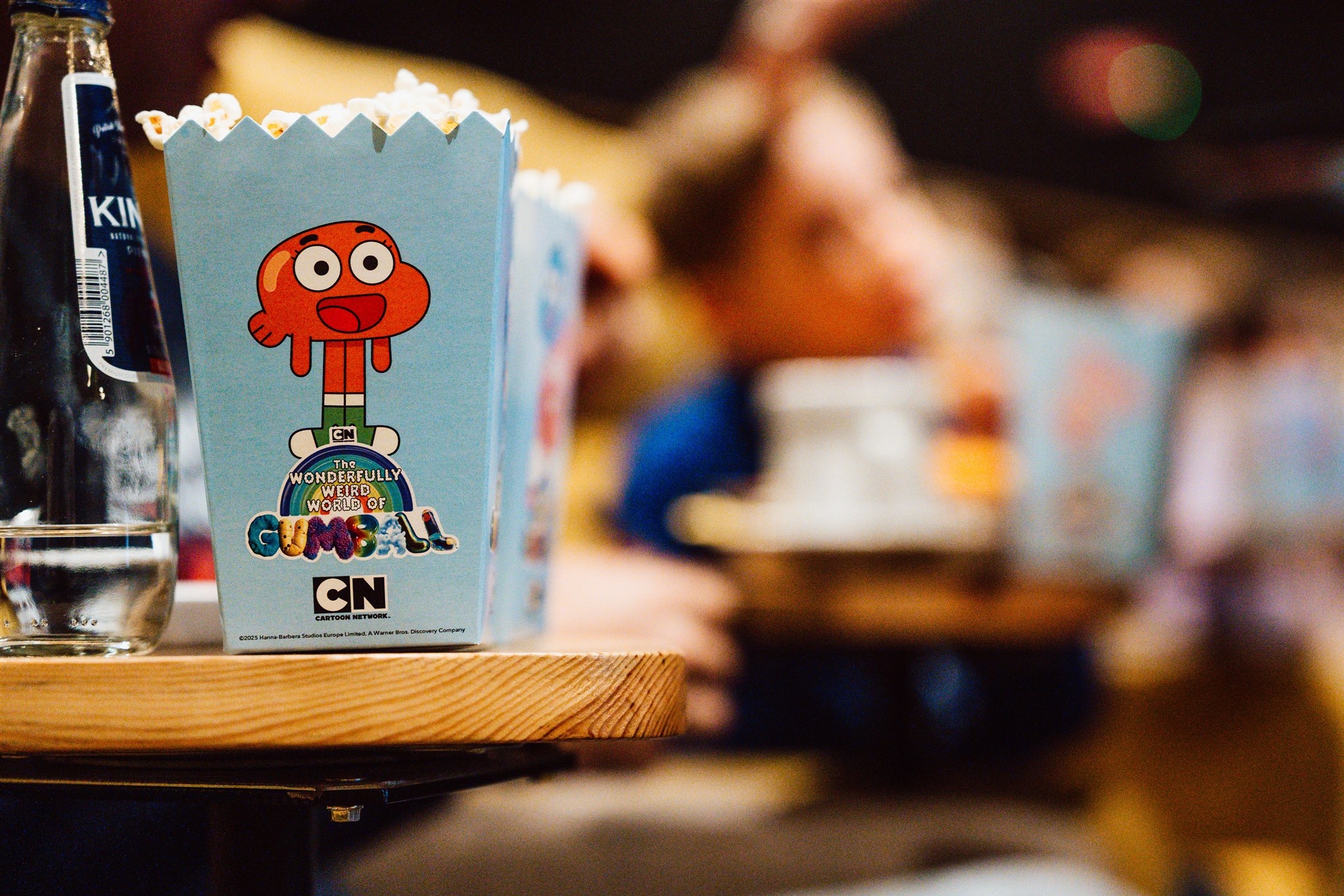 Kartony popcornu z motywem 'The Amazing World of Gumball' stoją na drewnianym blacie, obok butelka wody. W tle rozmyte postacie, tworząc klimat eventu dla dzieci.