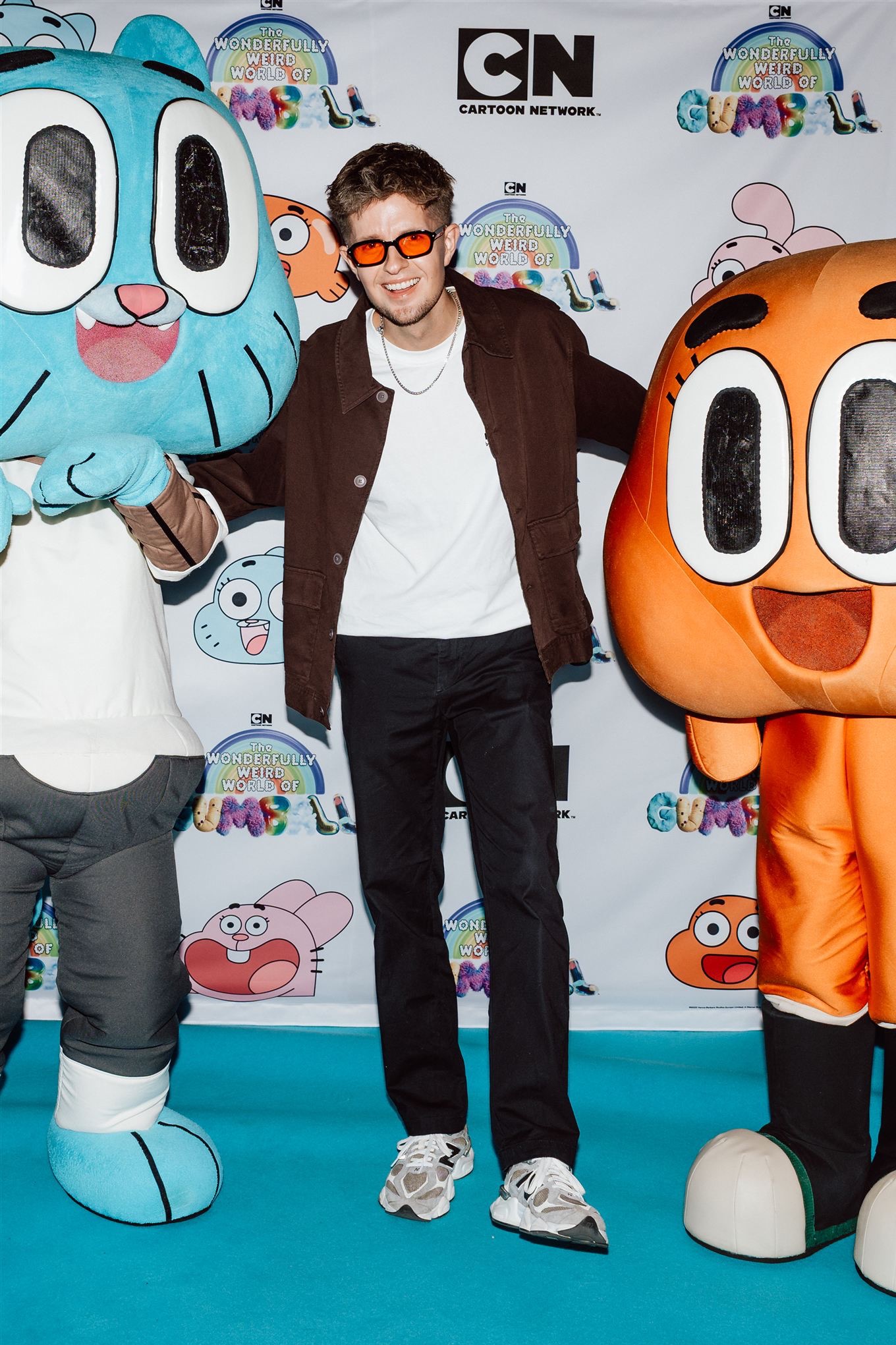 Mężczyzna w okularach pozuje z maskotkami Gumballa na tle logotypu Cartoon Network. Uśmiechnięty, swobodny strój, jasne tło z motywami z kreskówki.