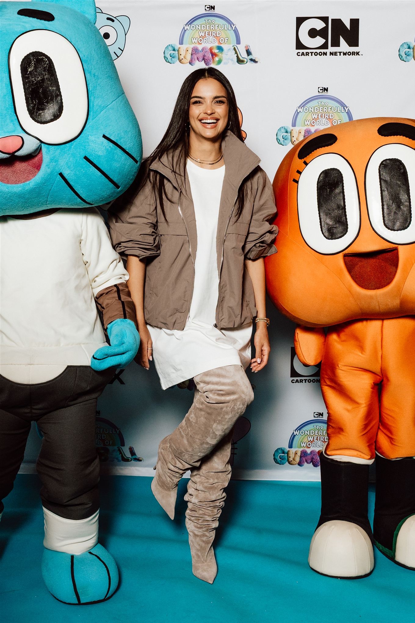 Uśmiechnięta kobieta w jasnej sukience i kurtce pozuje z maskotkami Gumballa na tle logotypu Cartoon Network, na evencie promocyjnym.