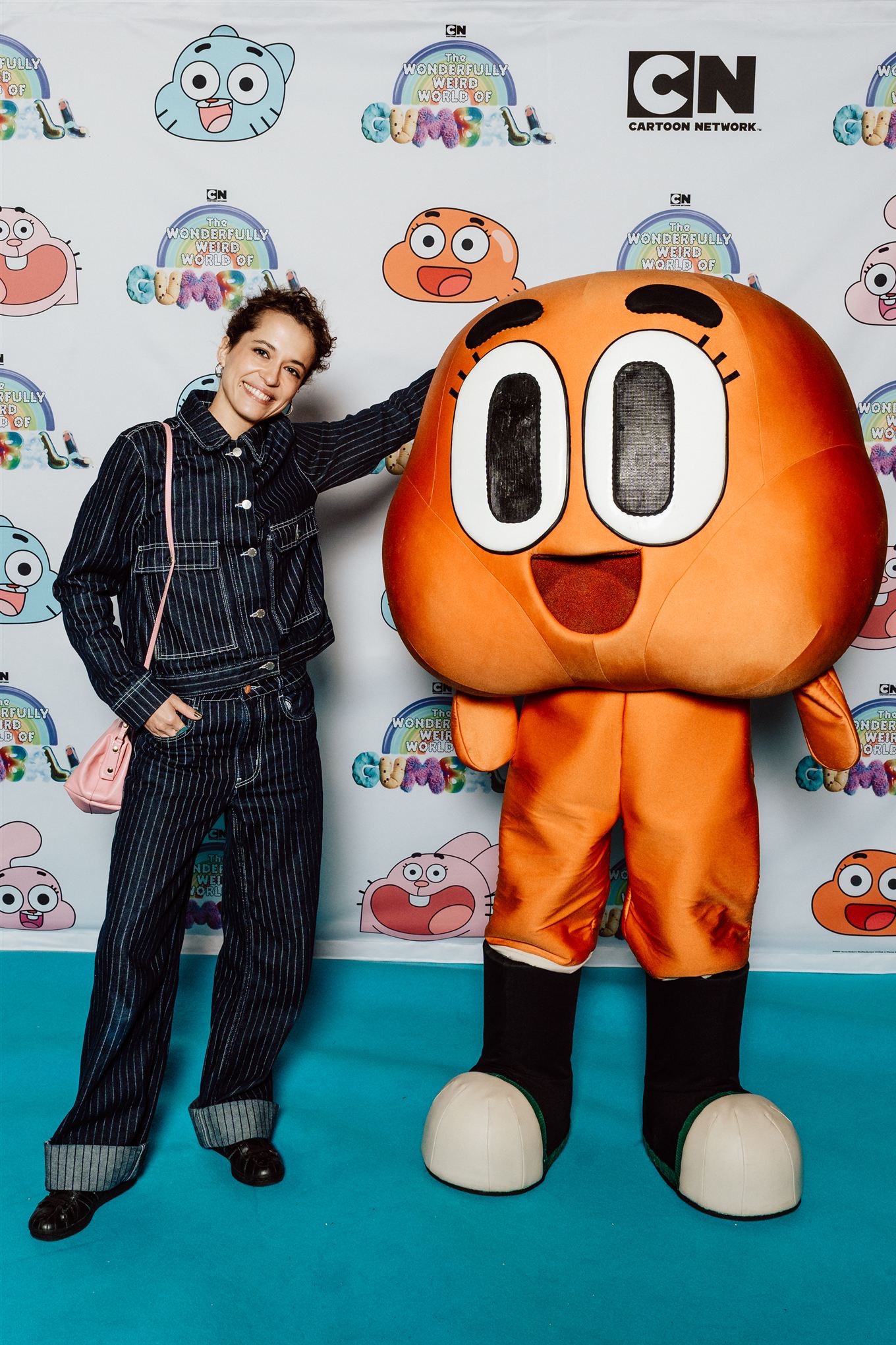 Kobieta w dżinsowym komplecie pozuje z animowaną postacią Darwin z 'The Amazing World of Gumball' na tle z logo Cartoon Network i motywami z serialu.