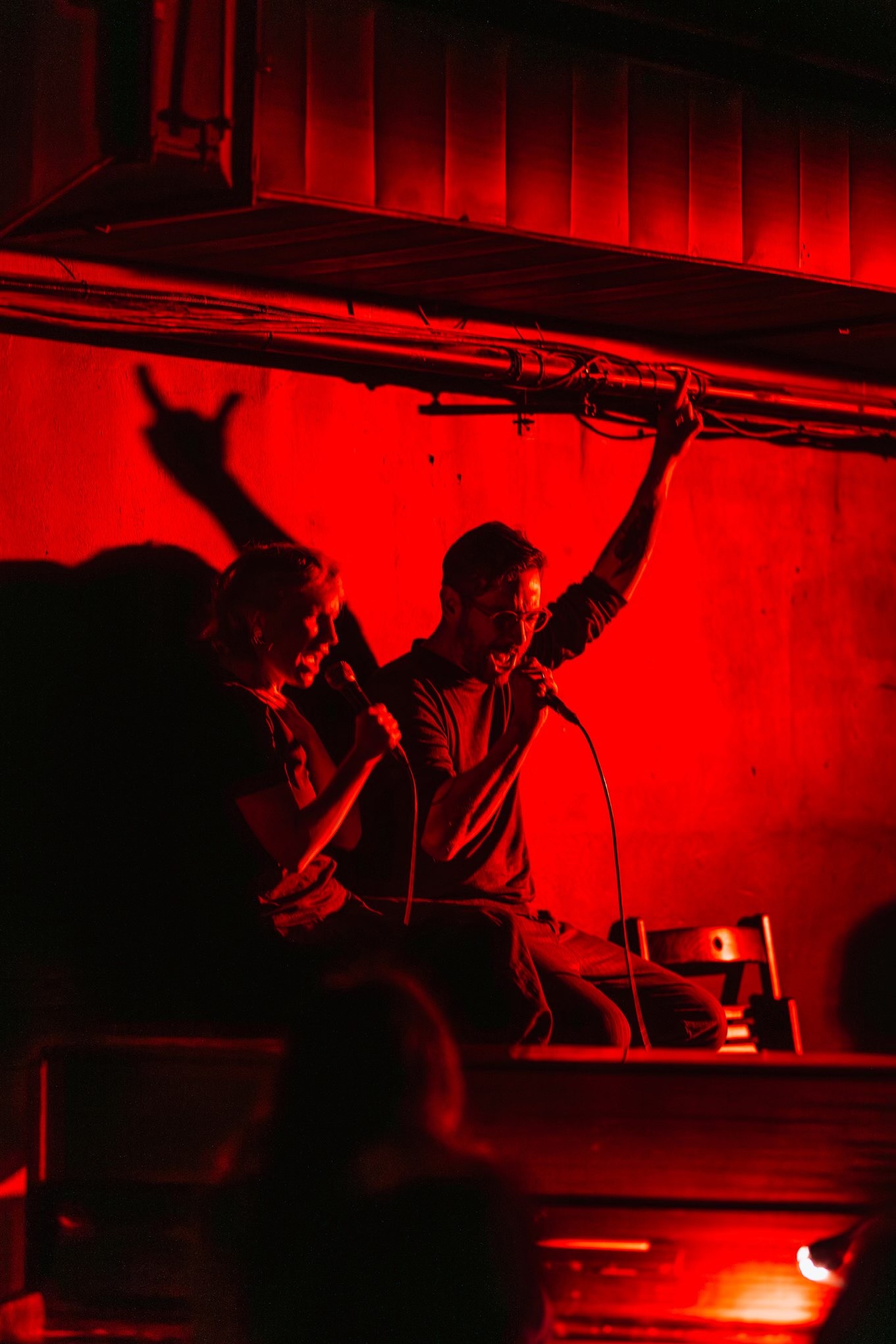 Duet wokalny na scenie, oświetlony intensywnym czerwonym światłem. Wokalistka z mikrofonem, wokalista z podniesioną ręką, ekspresyjne emocje, cień na ścianie.