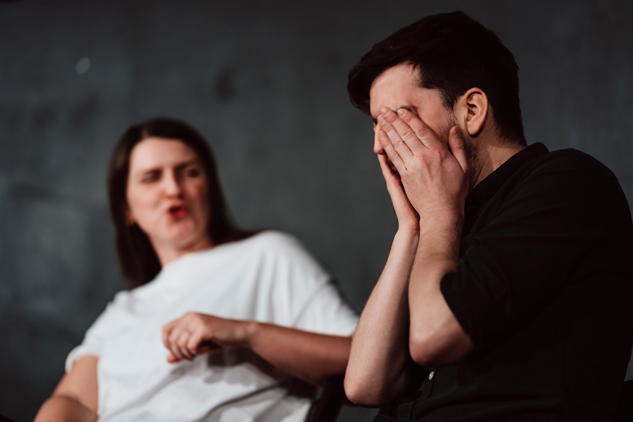 Mężczyzna zakrywa twarz dłońmi, siedząc obok kobiety z otwartymi ustami na rozmytym tle. Ujęcie z sesji biznesowej, ukazujące emocje i interakcje.