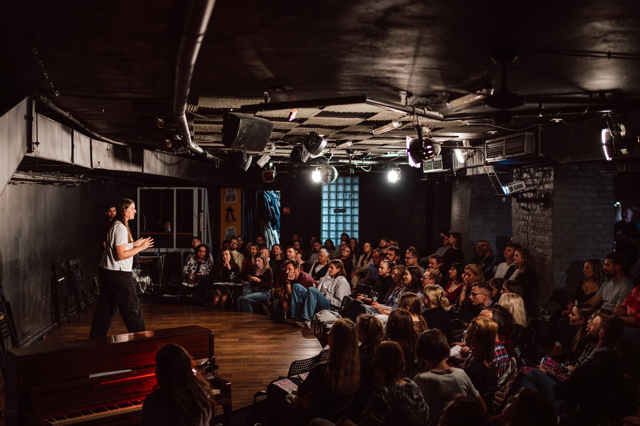 Kobieta przemawia na scenie przed publicznością w ciemnym wnętrzu z oświetleniem punktowym i pianinem. Ujęcie z widowni, skupienie na mówczyni.