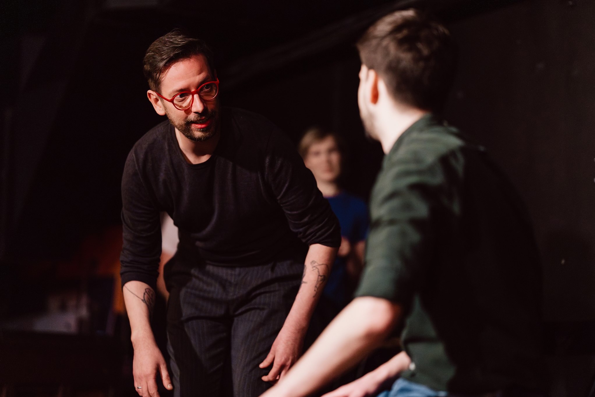 Dwóch mężczyzn w trakcie rozmowy na ciemnym tle. Jeden z nich ma czerwone okulary i tatuaż na ramieniu. Ujęcie z bliska, podkreślające interakcję.