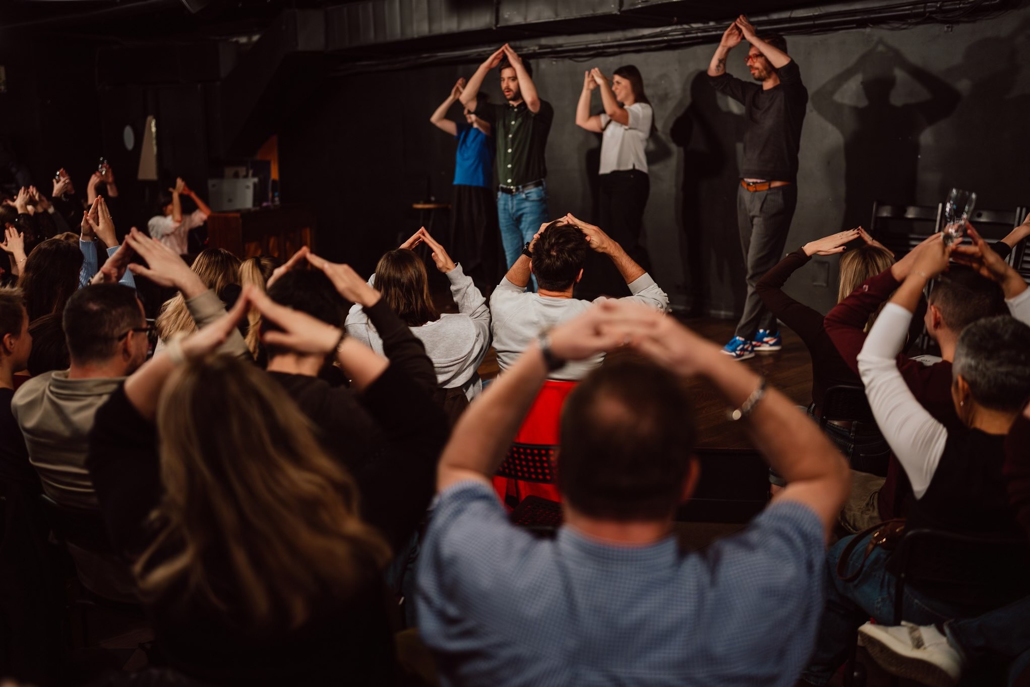 Widok zza pleców publiczności na scenę, gdzie grupa osób wykonuje gest dłońmi nad głową, tworząc trójkąt, w ciemnym wnętrzu klubu podczas występu.