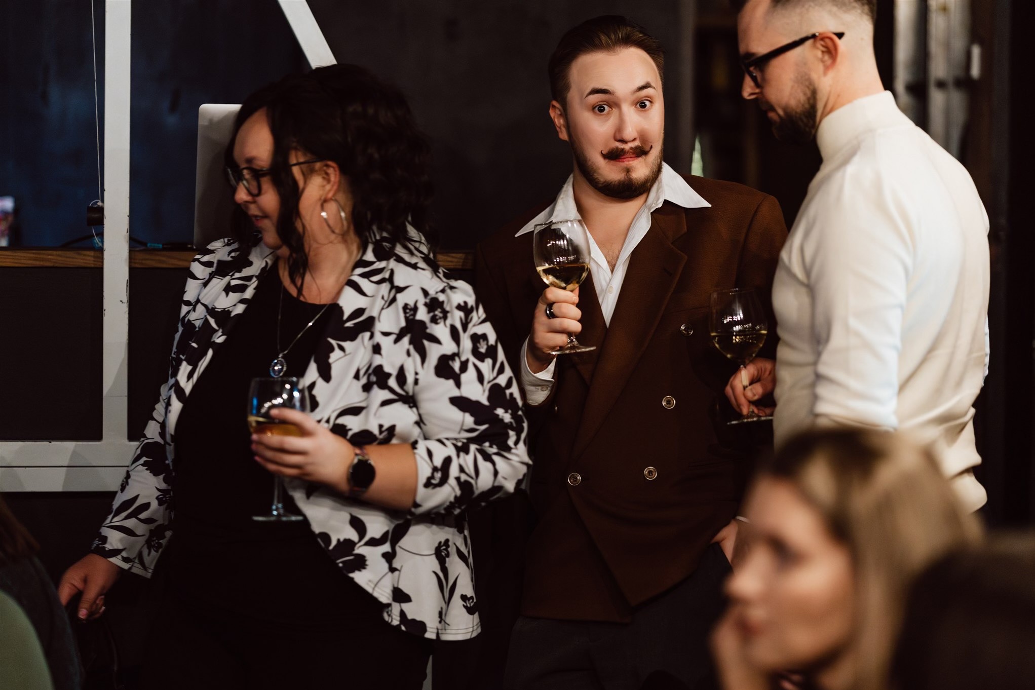 Elegancko ubrani goście z kieliszkami wina na evencie. Mężczyzna z wąsami w brązowej marynarce patrzy w obiektyw. Kobieta w kwiecistej marynarce i mężczyzna w bieli obok.