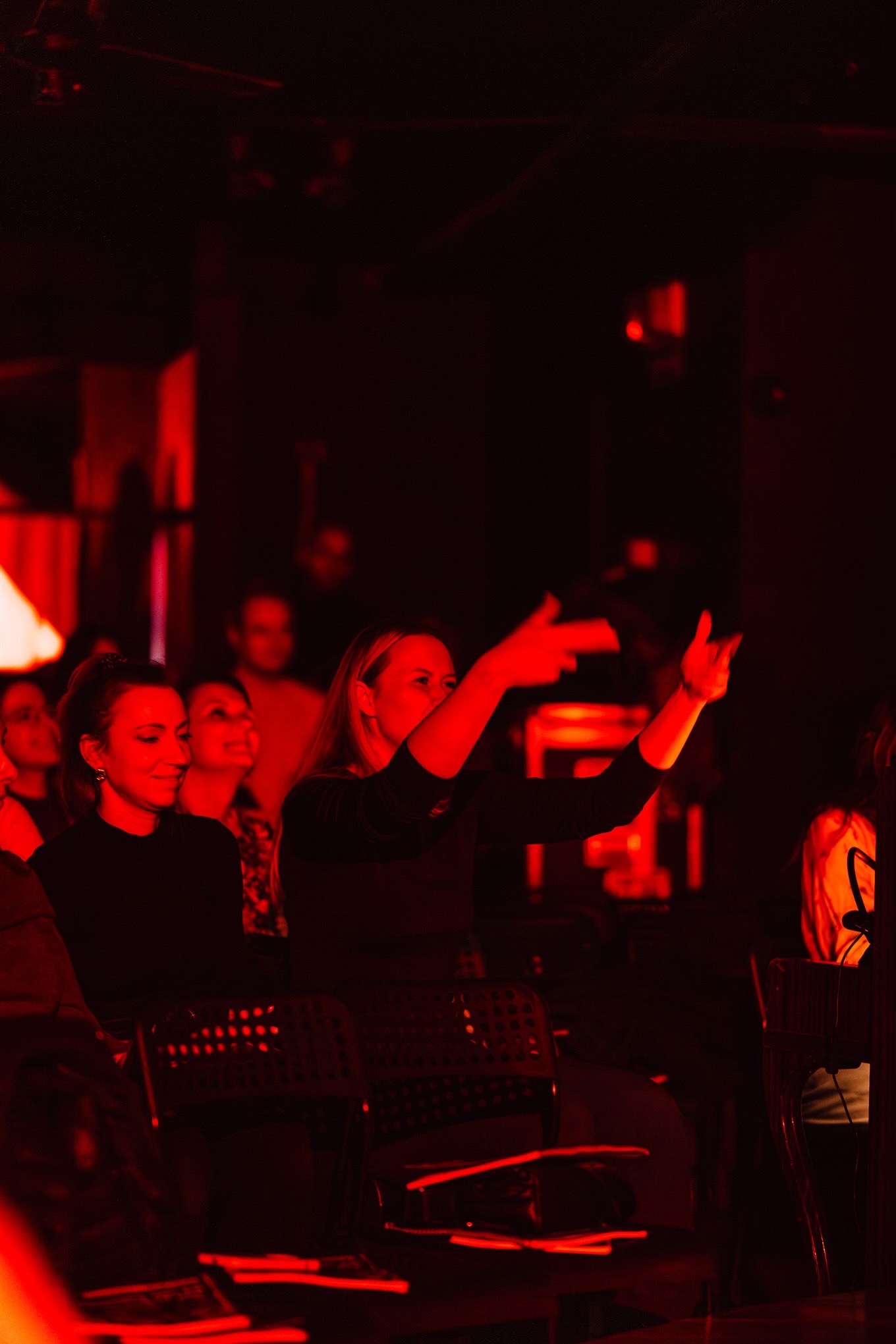Widownia w czerwonym oświetleniu, kobieta w czarnej bluzce gestykuluje, w tle siedzące osoby, metalowe krzesła z otworami, atmosfera koncertu lub występu.