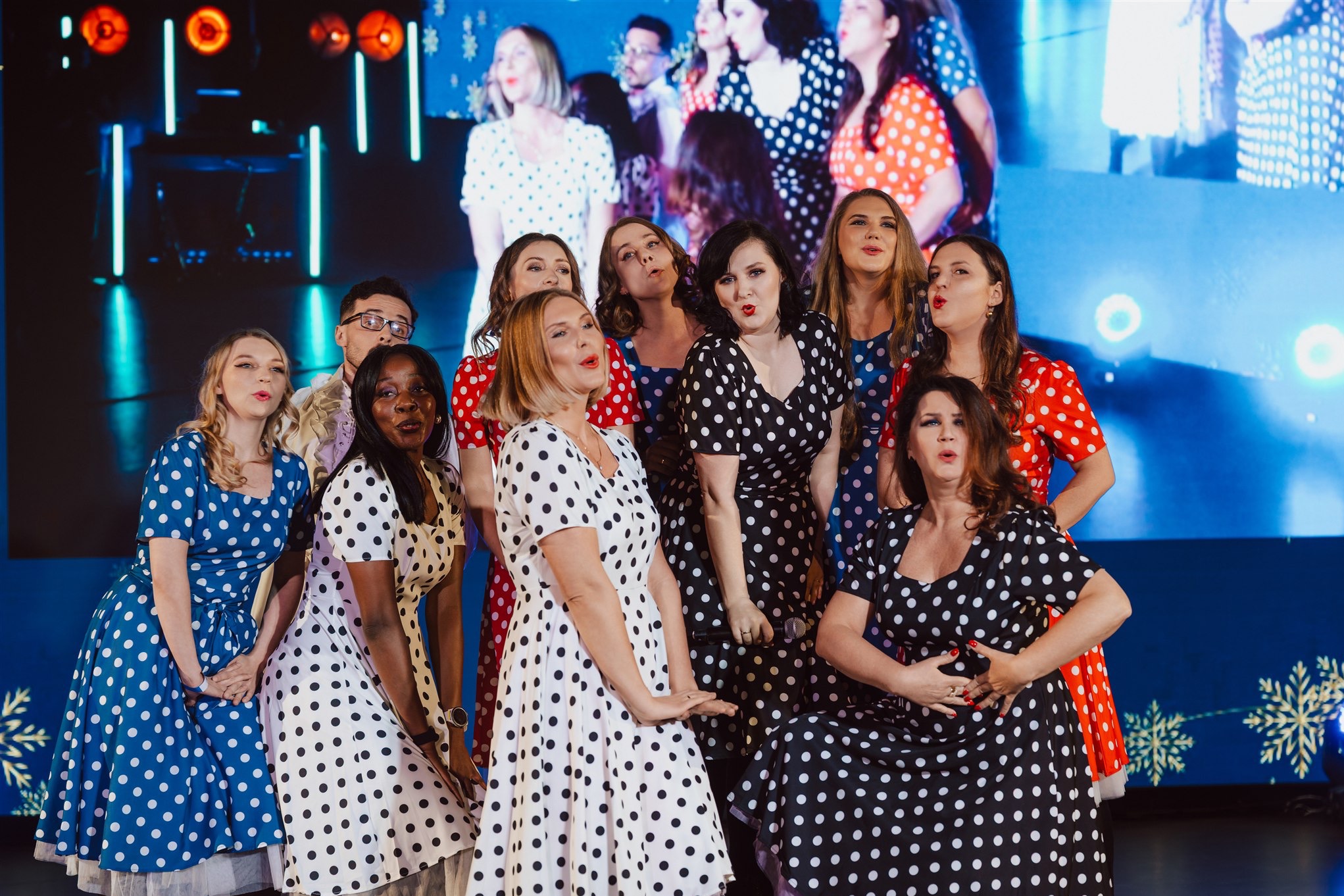 Zespół wokalny w kolorowych sukienkach w grochy, na scenie z niebieskim tłem i dekoracjami śnieżynkowymi, w trakcie występu.