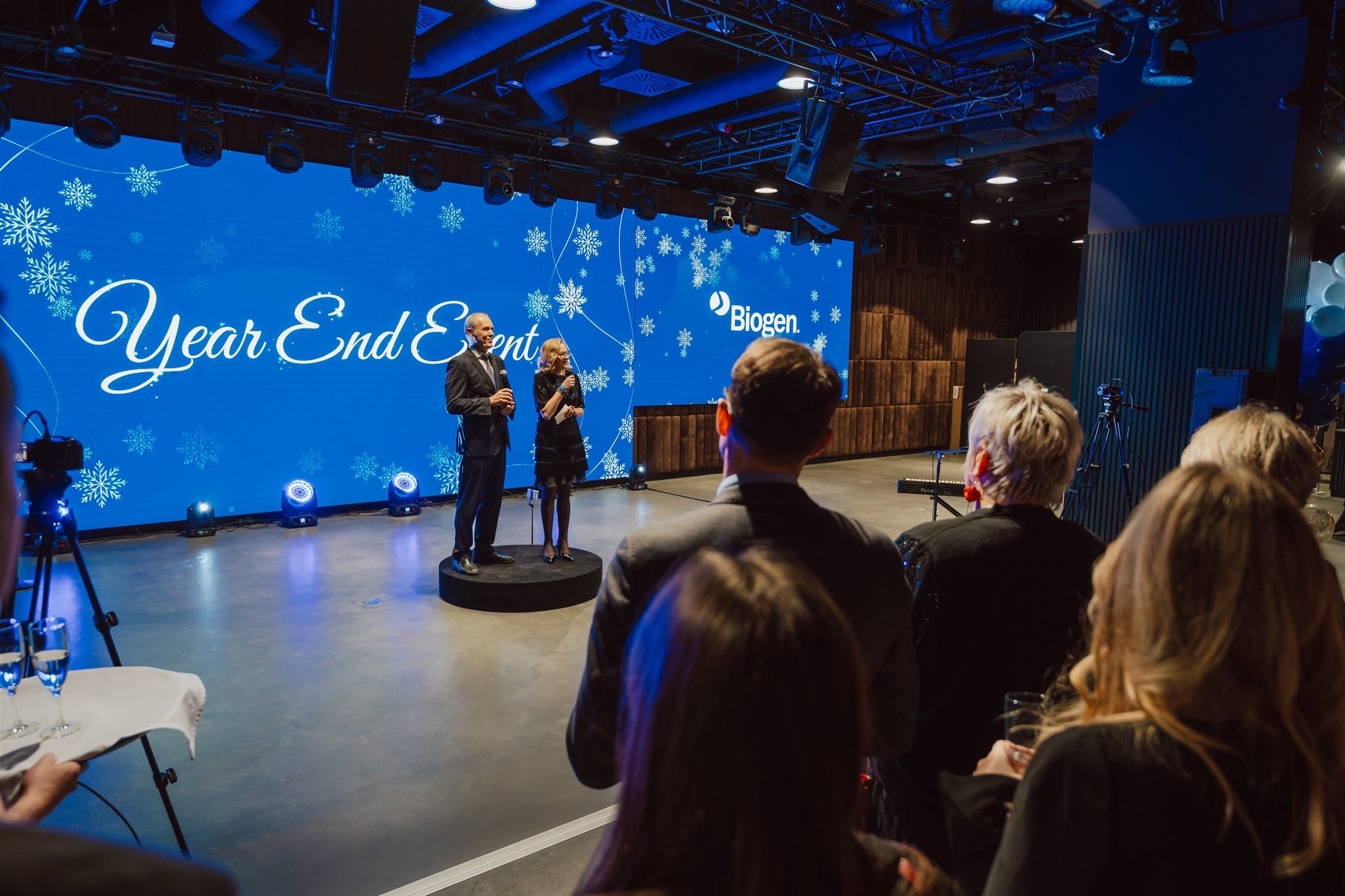 Prezenterzy na scenie podczas eventu firmowego 'Year End Event' z logo Biogen na ekranie. Widzowie obserwują, w tle oświetlenie sceniczne i dekoracje.