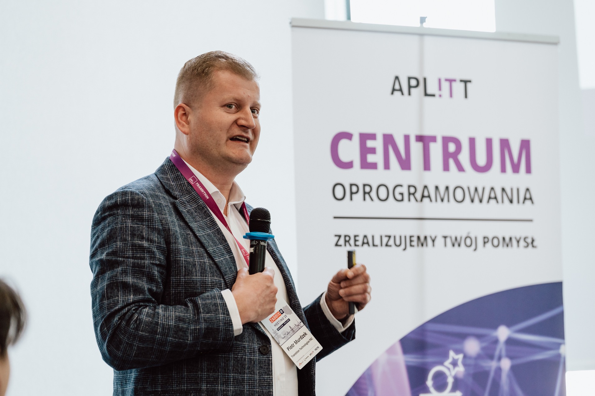 Mężczyzna w marynarce prezentuje na tle baneru APLITT Centrum Oprogramowania, trzymając mikrofon i wskaźnik. Ujęcie z konferencji, jasne tło.