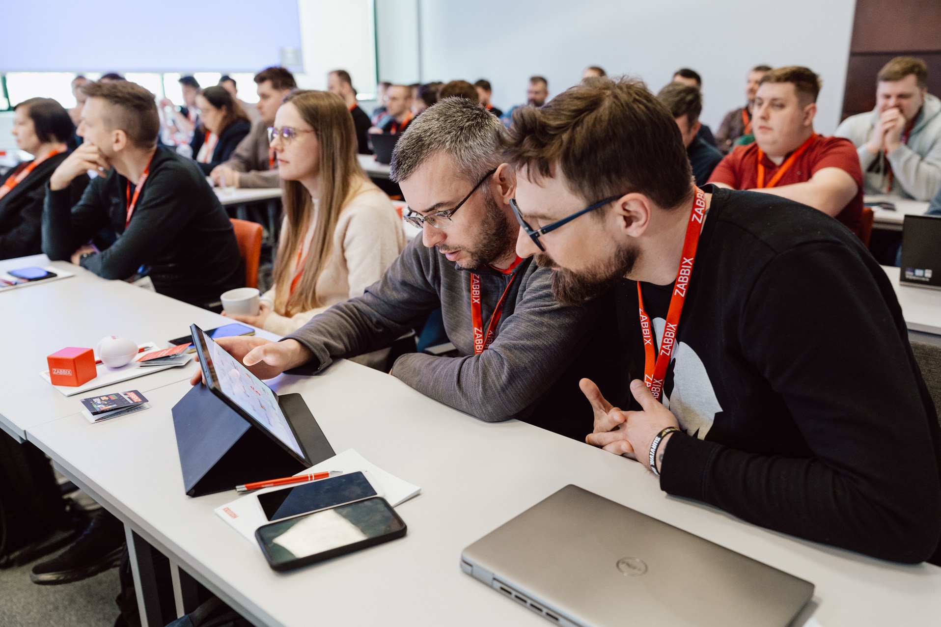 Konferencja Zabbix: uczestnicy skupieni na tablecie, dyskusja w trakcie prezentacji. Widoczne laptopy, telefony i materiały konferencyjne na białych stołach.