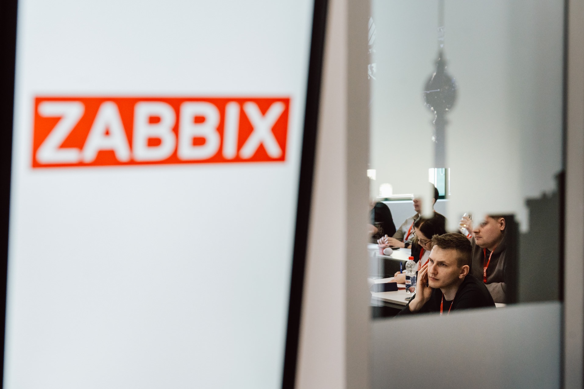 Konferencja Zabbix: Uczestnicy skupieni przy stole, widoczni przez szklaną ścianę z logo, w jasnym wnętrzu. Mężczyzna w czarnej koszulce w pierwszym planie.