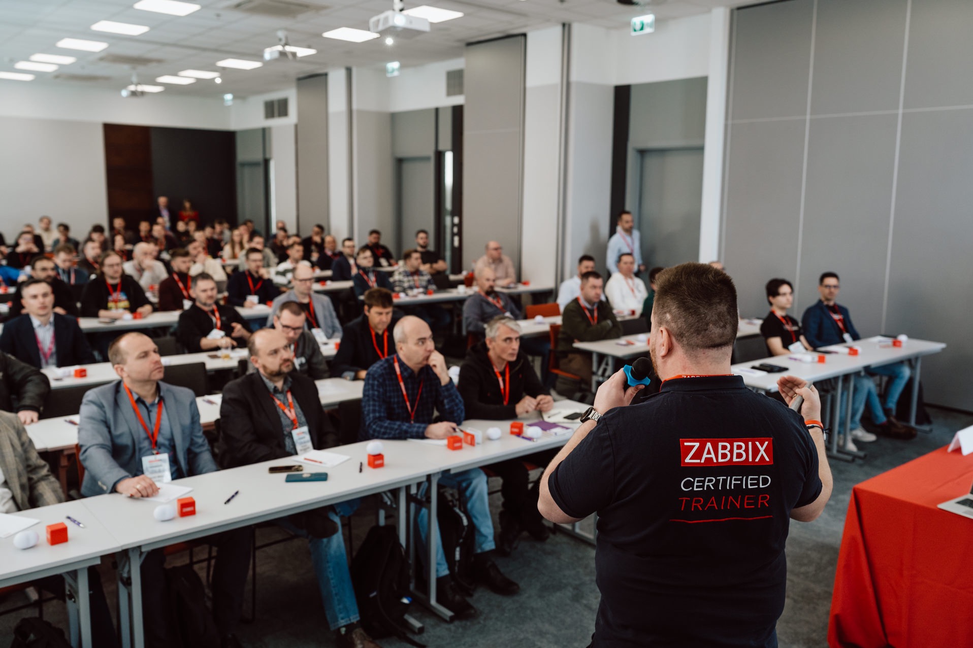 Prelegent z mikrofonem prowadzi szkolenie Zabbix dla grupy osób siedzących przy stołach w sali konferencyjnej. Widoczny tył koszulki trenera z logo Zabbix.