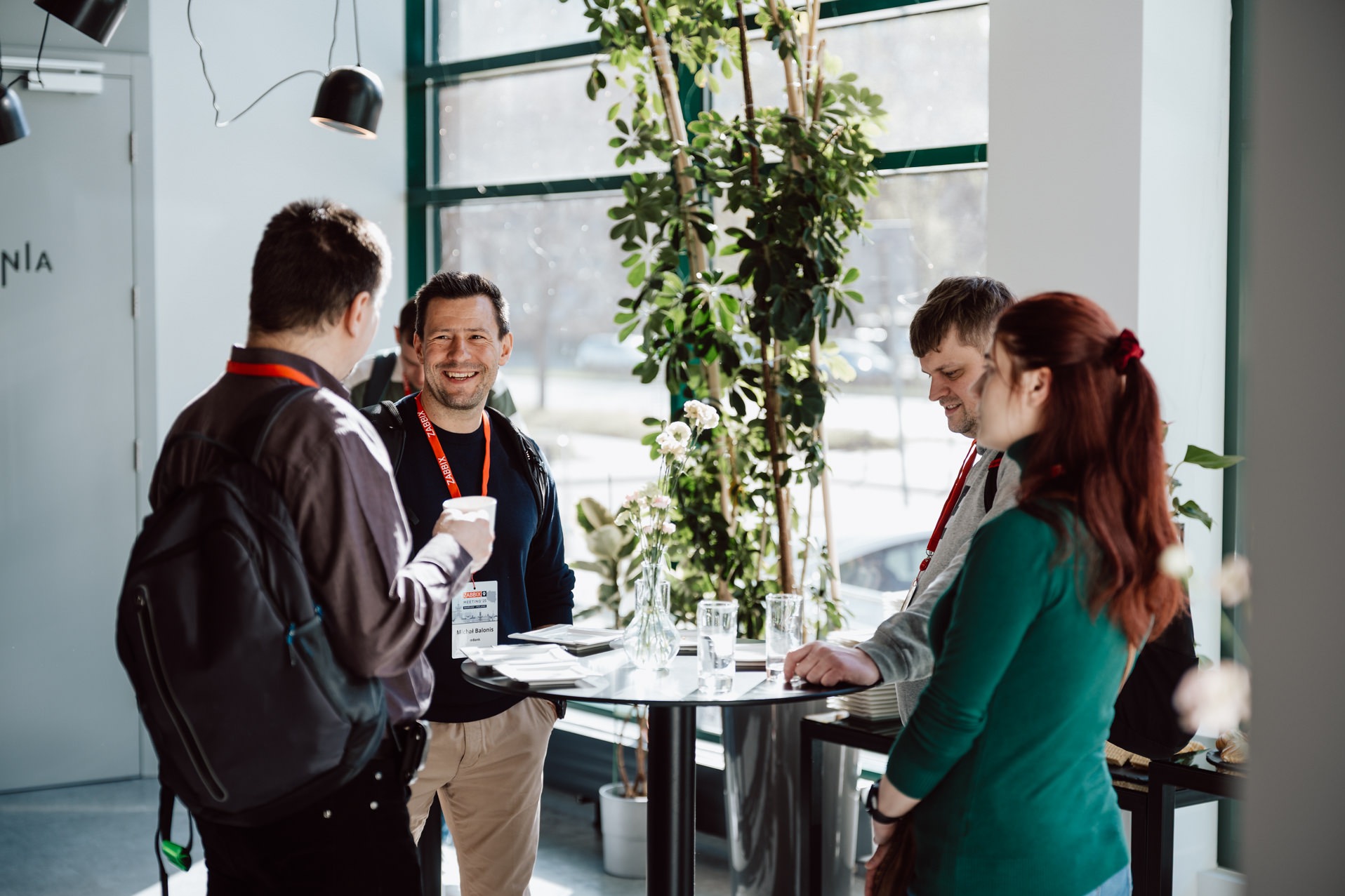 Uczestnicy konferencji w kuluarach: mężczyzna z identyfikatorem Zabbix uśmiecha się do rozmówcy, w tle jasne wnętrze z roślinami i dużym oknem.
