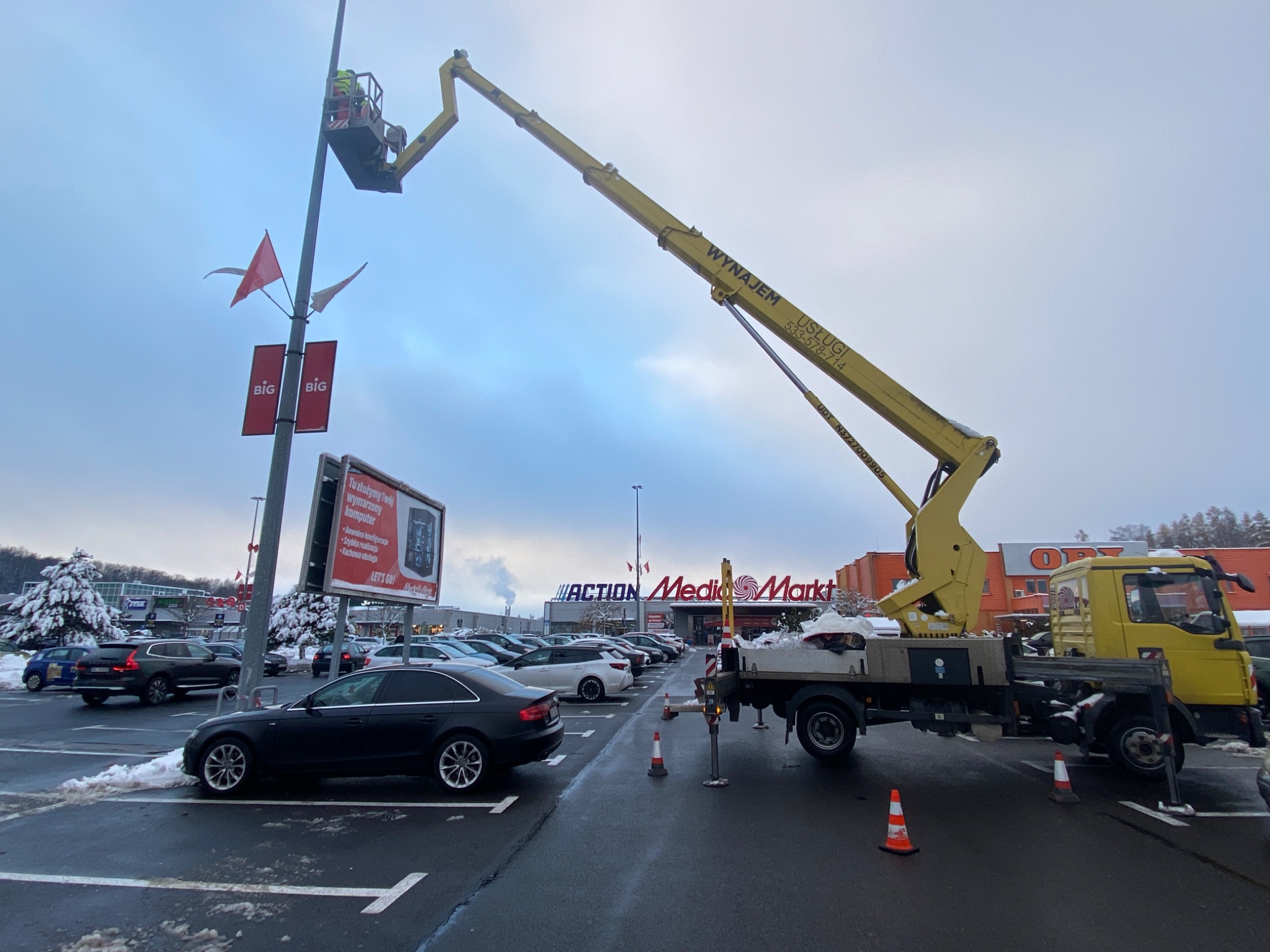 Żółty podnośnik koszowy na parkingu przed sklepem Action Media Markt. Pracownik w koszu przy słupie oświetleniowym. Śnieg na ziemi, pochmurne niebo.
