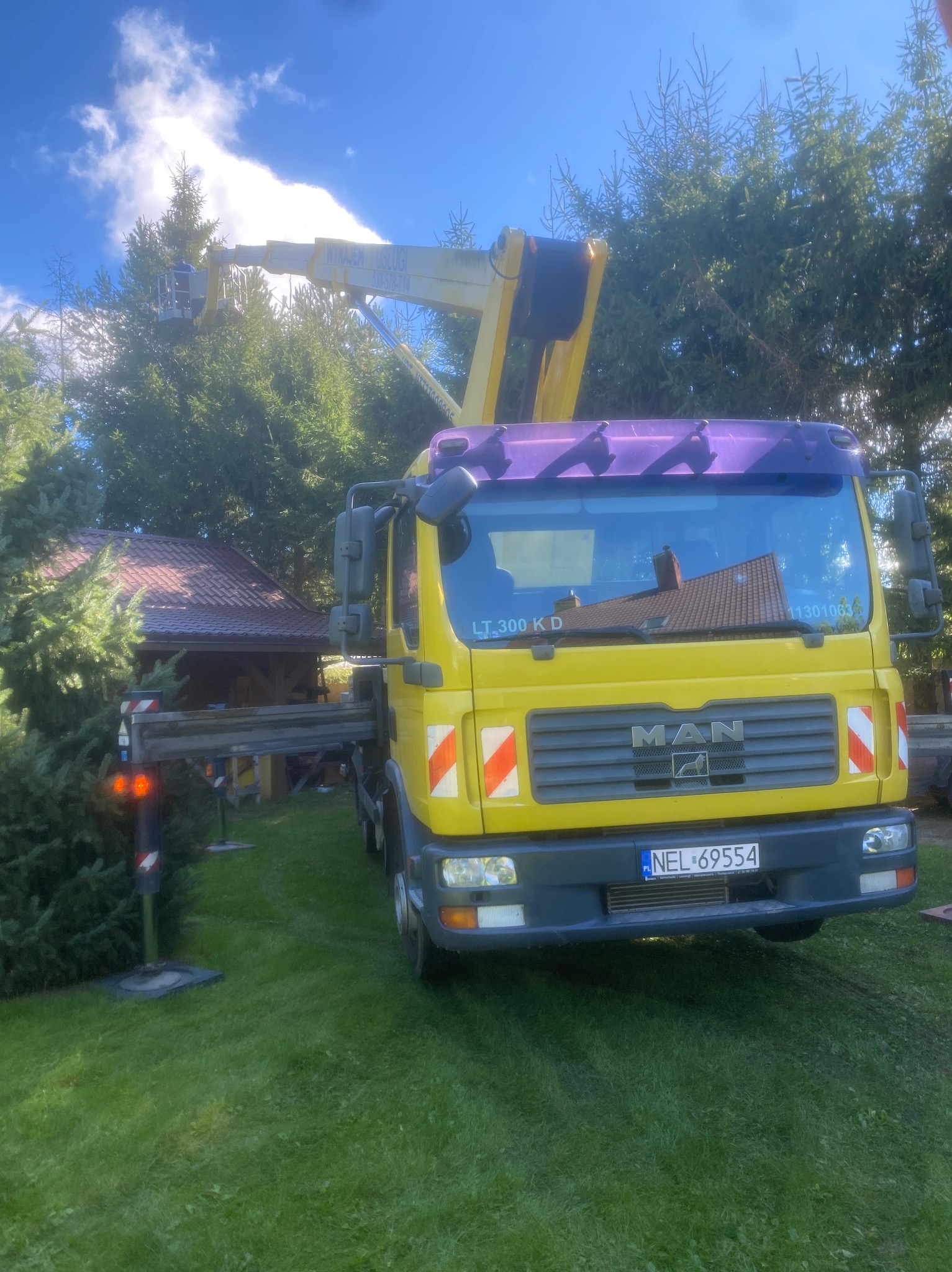 Żółty podnośnik koszowy MAN na trawniku, z wysięgnikiem sięgającym do korony drzewa. W tle widoczny budynek z czerwonym dachem. Błękitne niebo.