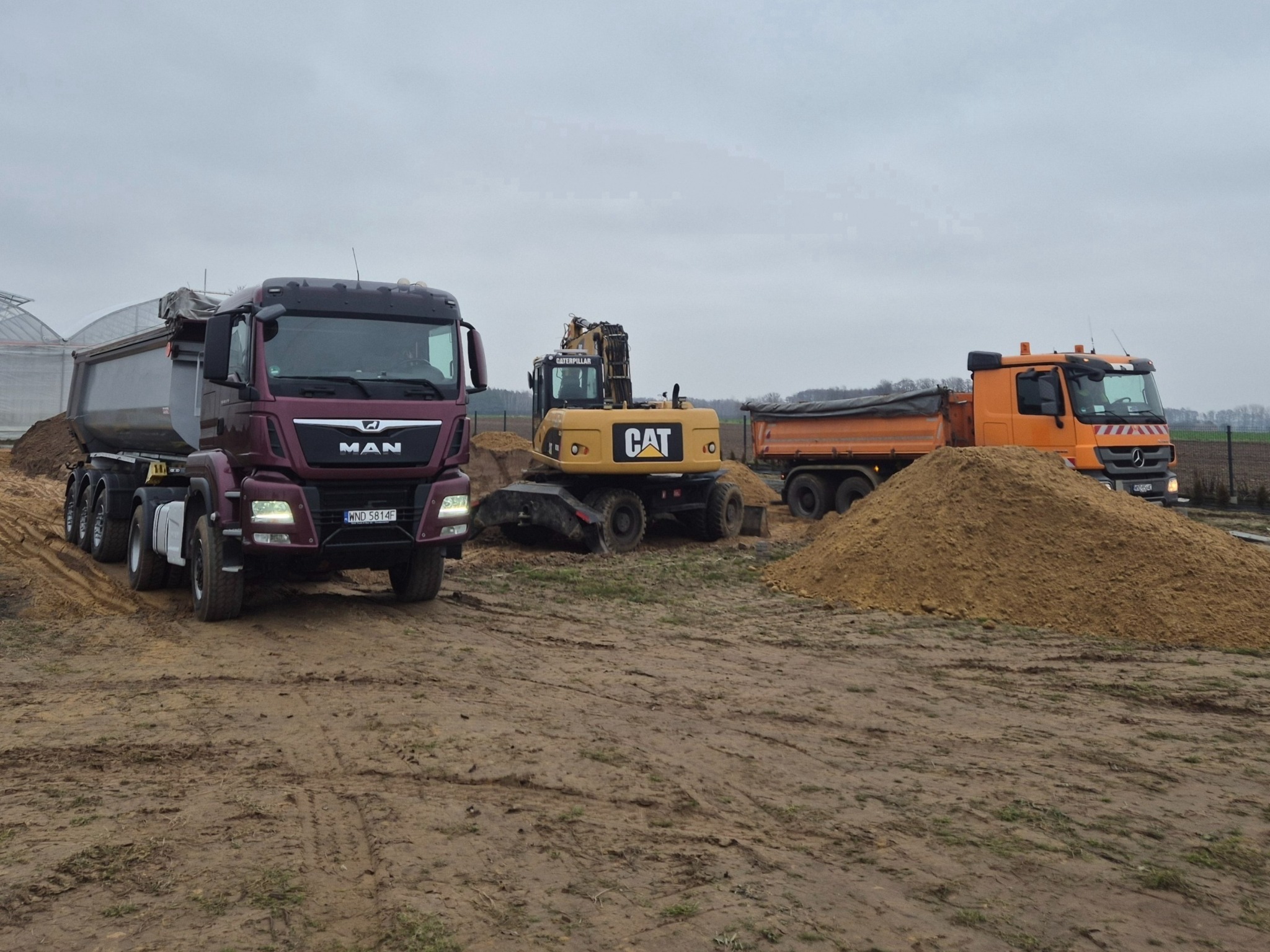 Ciężarówki MAN i Mercedes oraz koparka CAT na placu budowy, hałdy ziemi, szara pogoda, w tle szklarnia. Transport i prace ziemne.