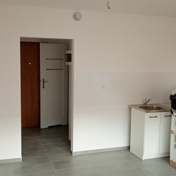 PEWNE SPRAWY SPÓŁKA Z OGRANICZONĄ ODPOWIEDZIALNOŚCIĄ - Wnętrze po remoncie: szare płytki podłogowe, białe ściany z panelami, biała szafka z umywalką i widok na drzwi. Minimalistyczny design, świeżo po wykończeniu.