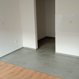 PEWNE SPRAWY SPÓŁKA Z OGRANICZONĄ ODPOWIEDZIALNOŚCIĄ - Wnętrze po remoncie: białe ściany, szare płytki podłogowe i fragment paneli. Widoczny otwór drzwiowy do kolejnego pomieszczenia. Wiadro malarskie w rogu kadru.