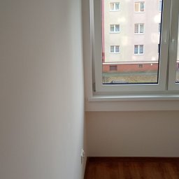 PEWNE SPRAWY SPÓŁKA Z OGRANICZONĄ ODPOWIEDZIALNOŚCIĄ - Jasne, świeżo pomalowane wnętrze pokoju z drewnianą podłogą. Widok z okna na blok mieszkalny. Minimalistyczny wystrój po remoncie.