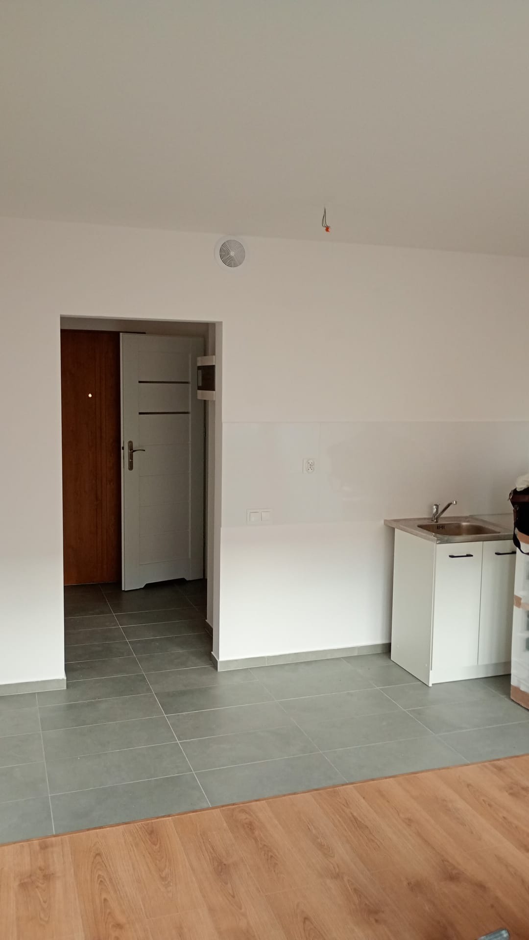 Wnętrze po remoncie: szare płytki podłogowe, białe ściany z panelami, biała szafka z umywalką i widok na drzwi. Minimalistyczny design, świeżo po wykończeniu.