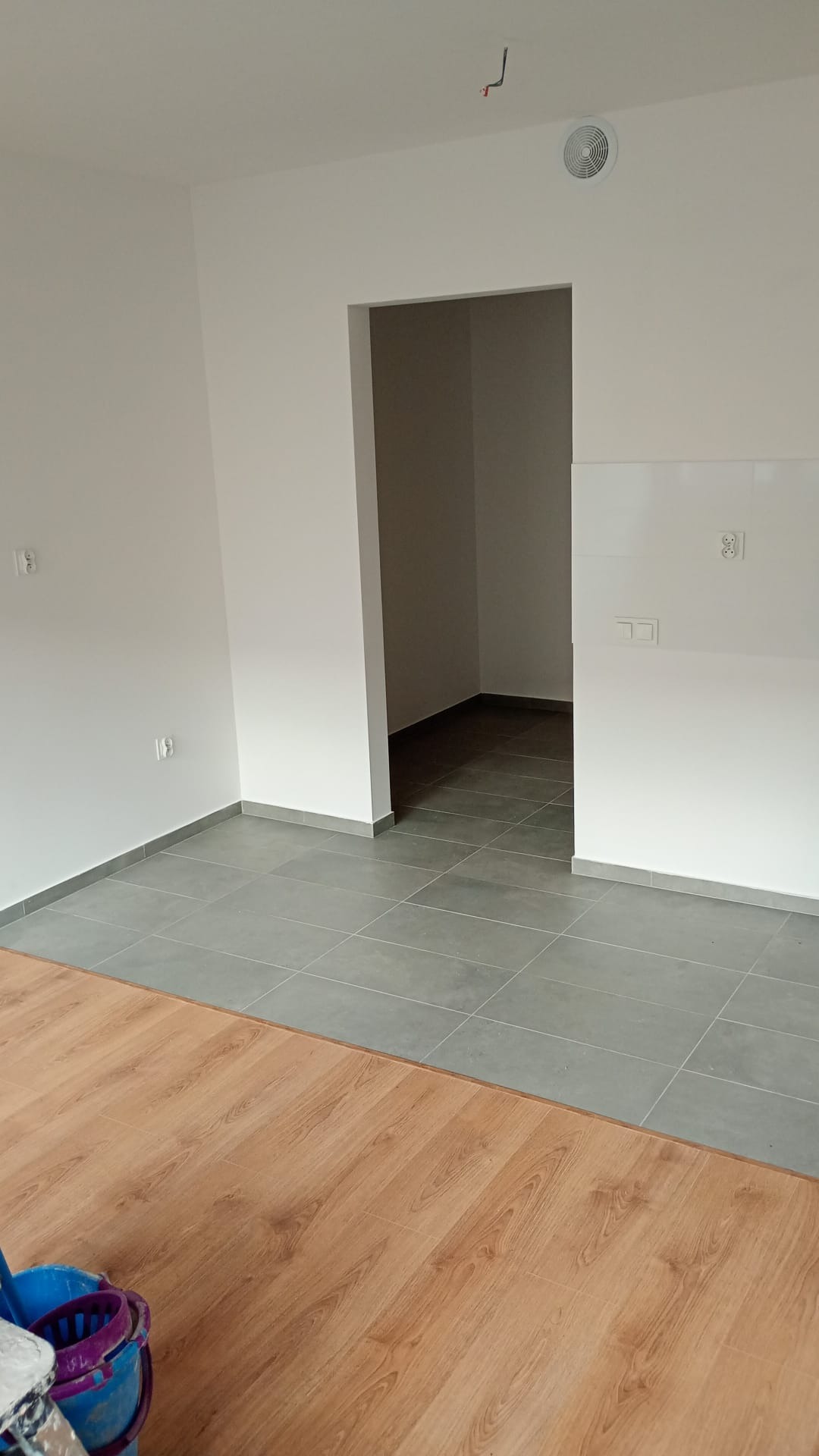 Wnętrze po remoncie: białe ściany, szare płytki podłogowe i fragment paneli. Widoczny otwór drzwiowy do kolejnego pomieszczenia. Wiadro malarskie w rogu kadru.