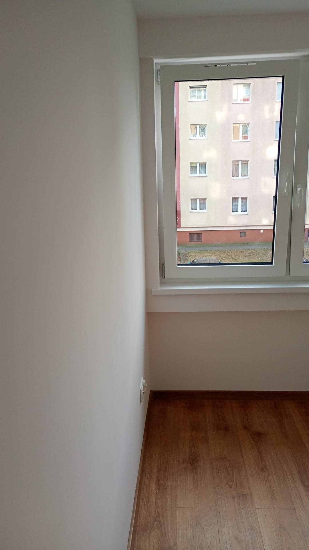 Jasne, świeżo pomalowane wnętrze pokoju z drewnianą podłogą. Widok z okna na blok mieszkalny. Minimalistyczny wystrój po remoncie.