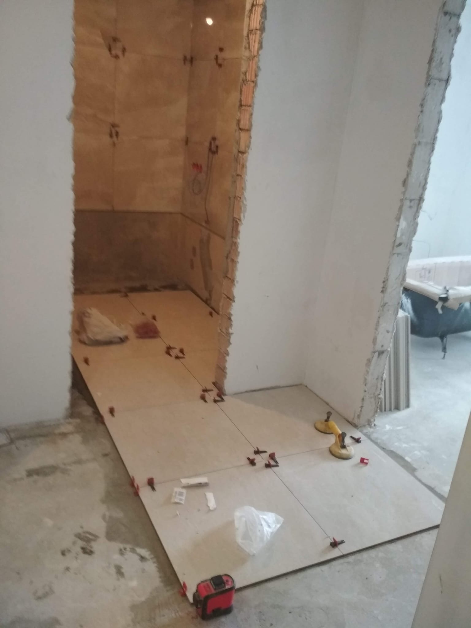Układanie płytek podłogowych w łazience w trakcie remontu. Widoczne przyssawki, poziomica laserowa i krzyżyki dystansowe. Surowy stan ścian i podłoża.