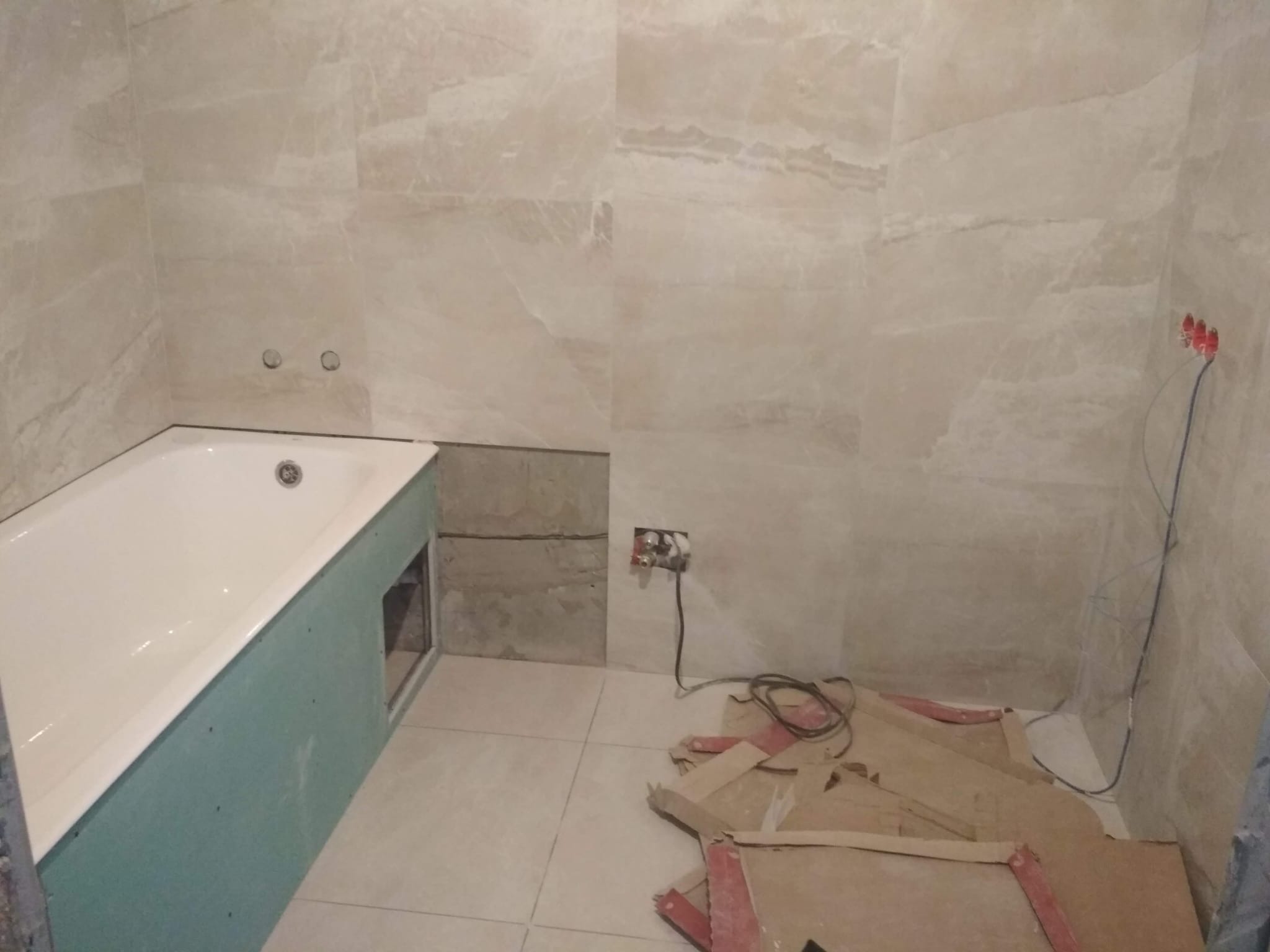 Remont łazienki w trakcie: obudowana wanna z zielonej płyty GK, częściowo położone płytki imitujące marmur, wystające kable i kartony na podłodze.