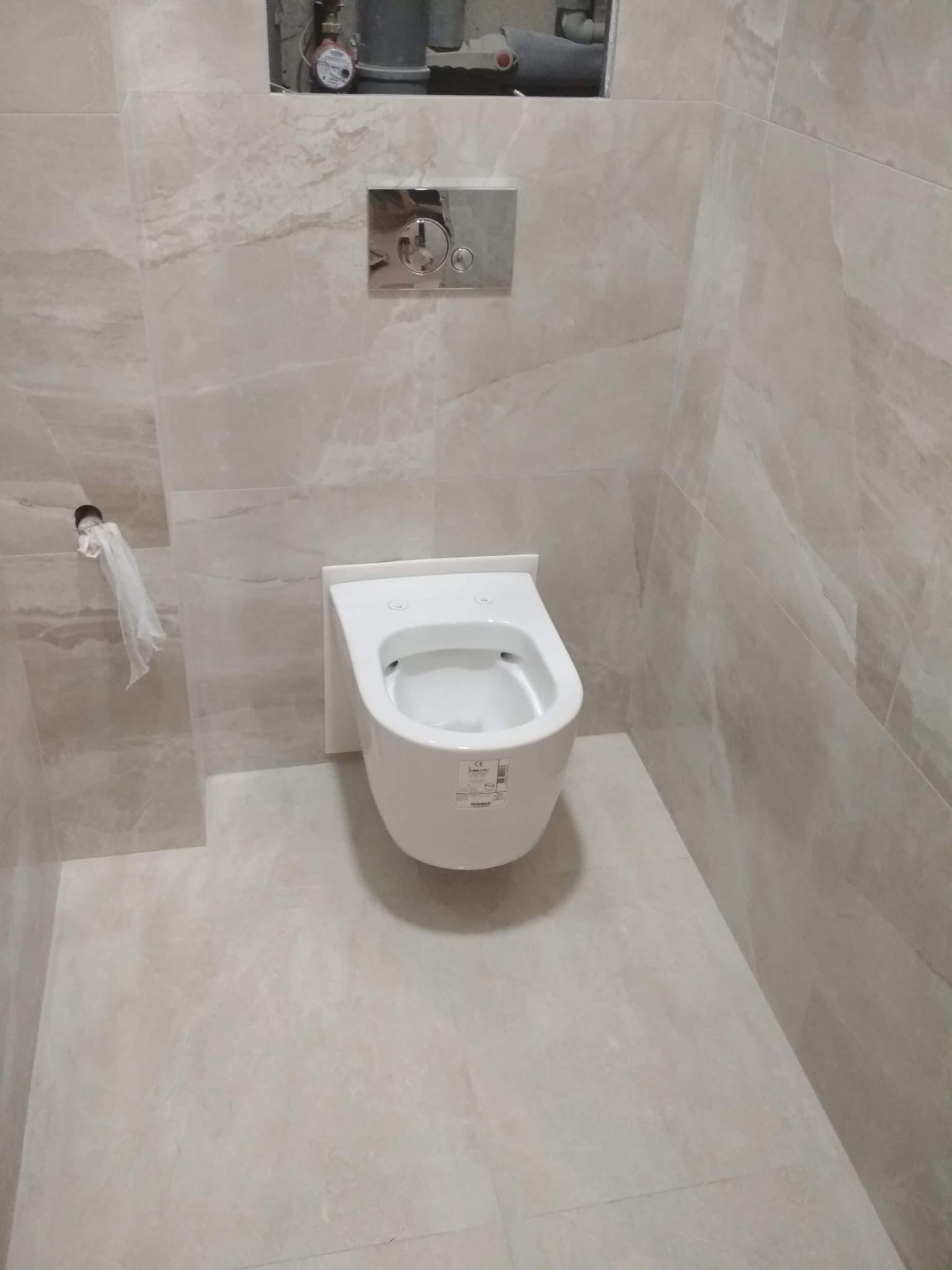 Zamontowana, podwieszana toaleta w wykończonej łazience z beżowymi płytkami imitującymi marmur. Widoczny chromowany przycisk spłuczki i fragment instalacji.