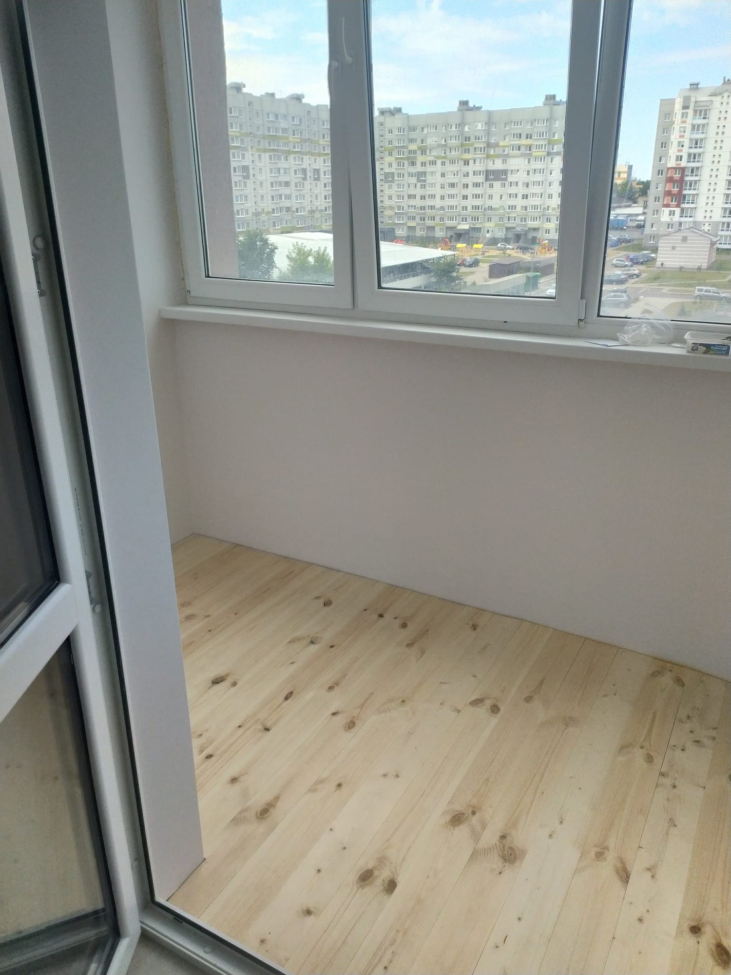 Jasne, drewniane deski na podłodze balkonu w Gdańsku, widok z otwartych drzwi balkonowych na budynki mieszkalne za oknem. Świeżo pomalowane ściany.