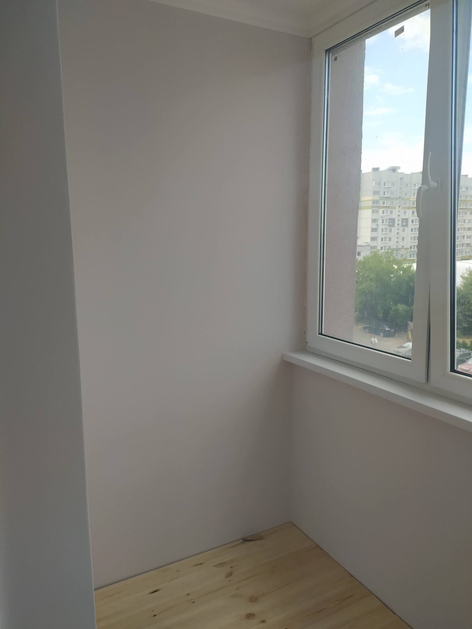 Jasny, odnowiony balkon z drewnianą podłogą i świeżo pomalowanymi ścianami w odcieniach beżu. Widok na miasto przez białe okno PCV. Minimalistyczny design.