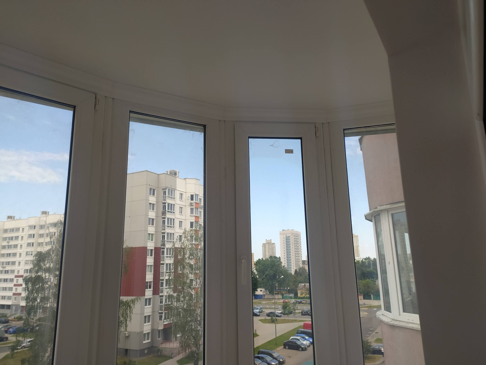 Zabudowany balkon z białymi ramami okiennymi, widok na osiedle miejskie w słoneczny dzień. Widoczne budynki mieszkalne i zieleń. Okna po remoncie, czyste szyby.