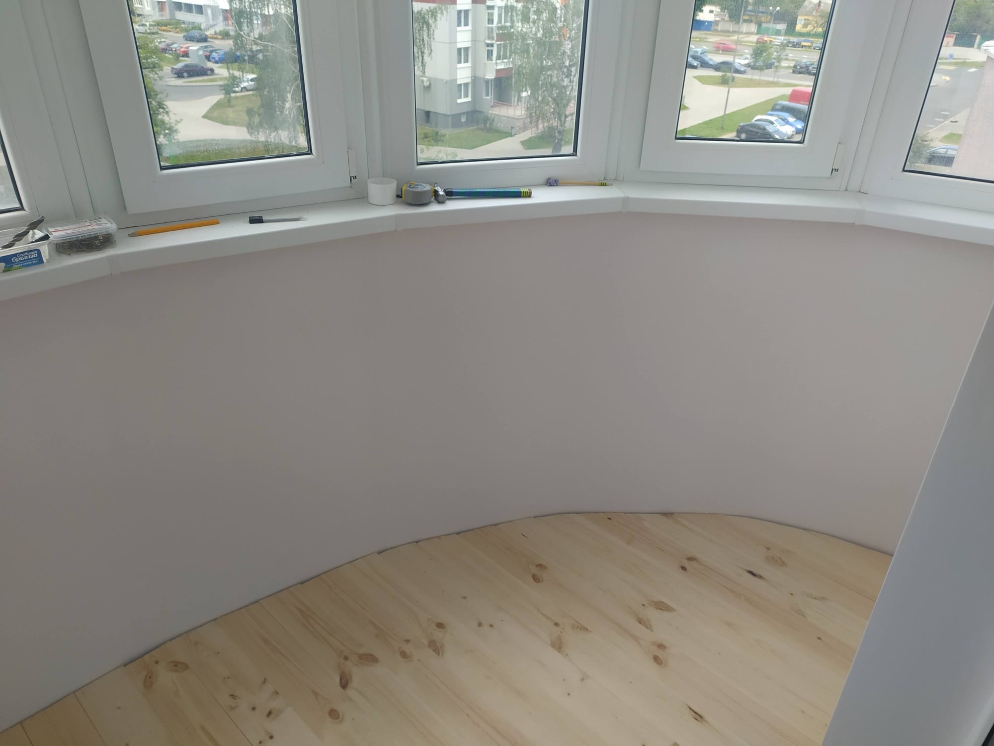 Półokrągły balkon po remoncie: jasna podłoga z desek, świeżo pomalowane ściany na delikatny róż, okna z widokiem na miasto. Na parapecie narzędzia.