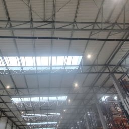 PPHU Elektromat Mateusz Musiała - Wnętrze hali magazynowej z widoczną konstrukcją dachu, oświetleniem i instalacjami pod stropem. Widoczne regały magazynowe i wózek widłowy.