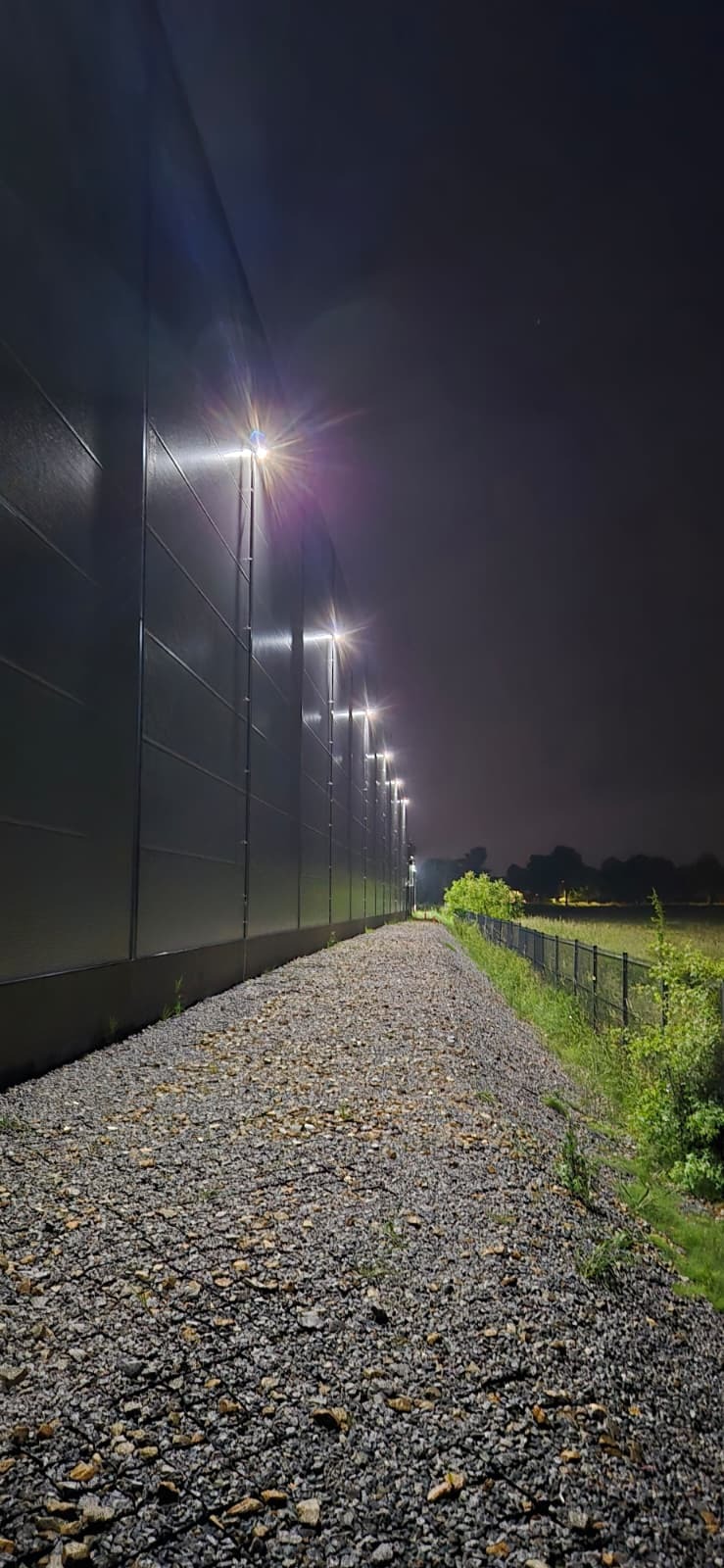 Nocne oświetlenie elewacji budynku przemysłowego z widocznymi lampami LED, oświetlającymi ścieżkę z kamieni. Widok z dołu, perspektywa zbieżna.