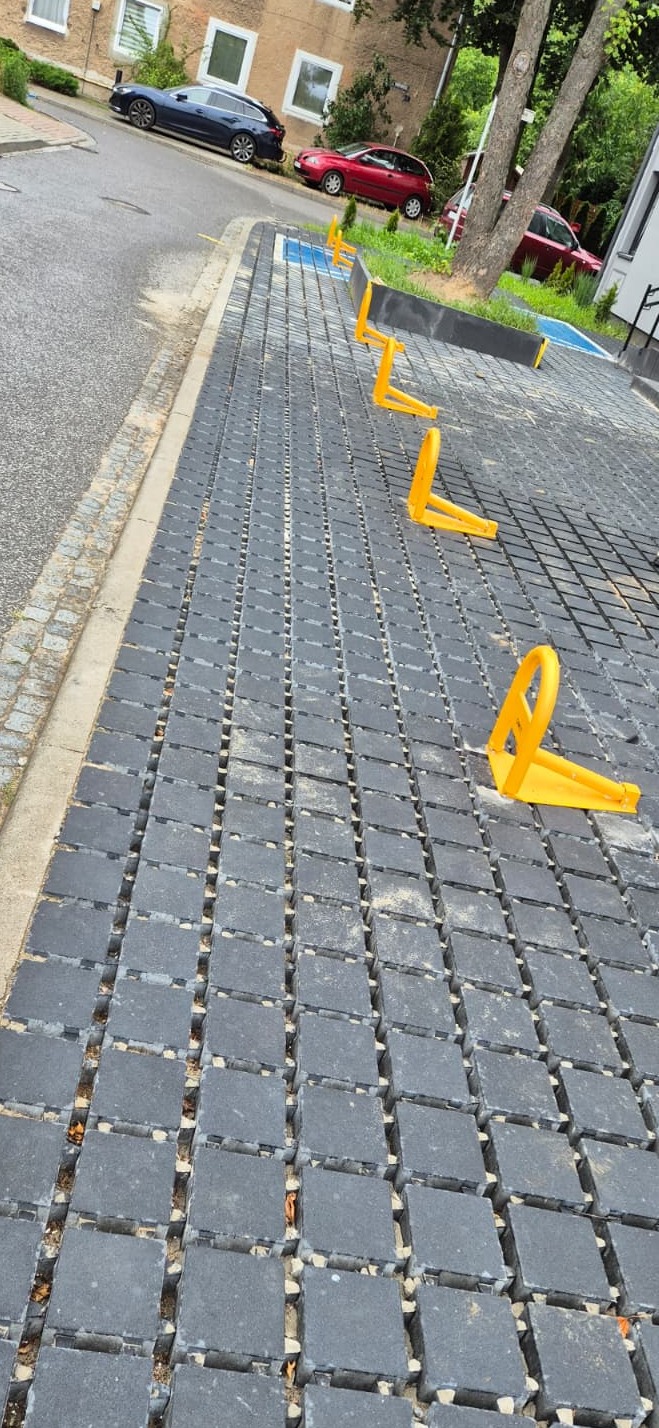 Szara nawierzchnia parkingu z ażurowych płyt i żółtymi blokadami miejsc, w tle budynek mieszkalny i zaparkowane auta. Ujęcie z perspektywy chodnika.