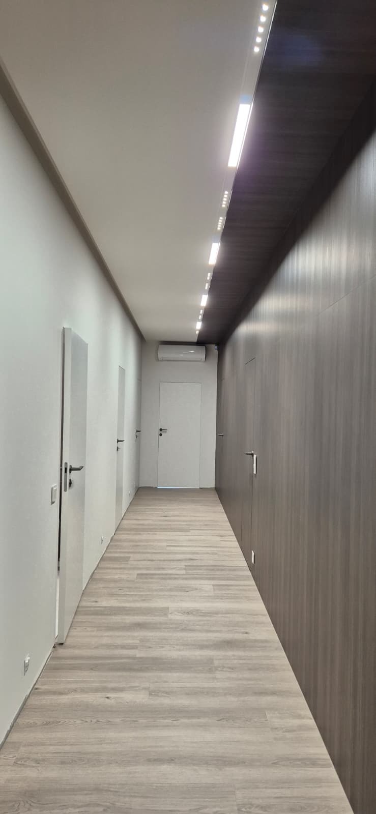 Długi korytarz z panelami podłogowymi i oświetleniem LED. Po lewej stronie białe drzwi, po prawej ciemne panele ścienne. Widoczny klimatyzator.