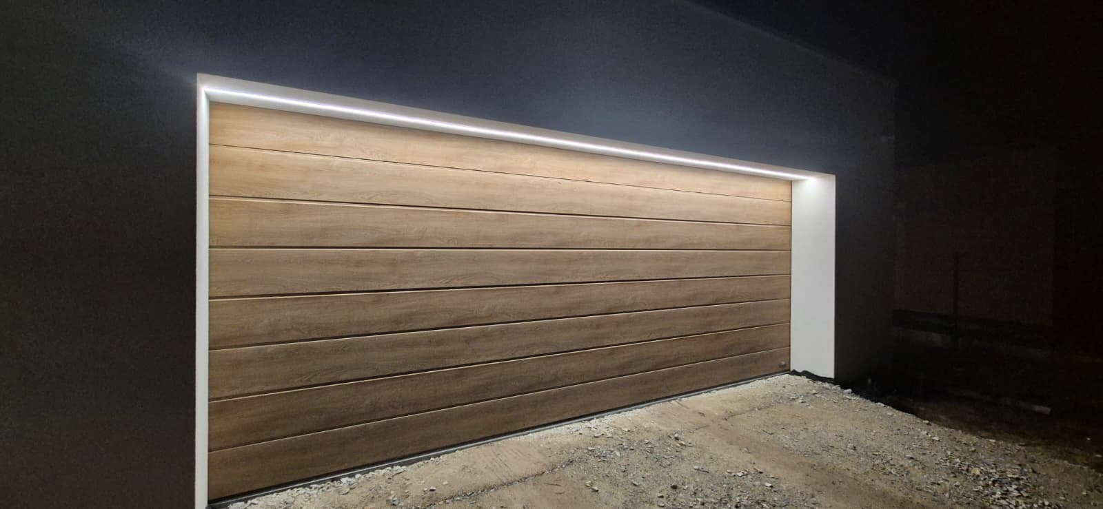 Drewniana brama garażowa segmentowa z subtelnym oświetleniem LED, widoczna z zewnątrz, na tle ciemnej elewacji. Nowoczesny design i precyzyjny montaż.