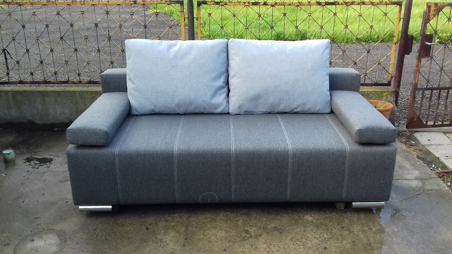 Szara, dwuosobowa sofa z poduszkami na zewnątrz, na tle metalowego ogrodzenia i zieleni. Minimalistyczny design, metalowe nóżki, proste linie. Widoczne ślady użytkowania na podłożu.