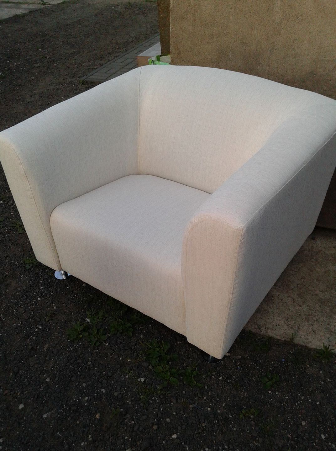 Kremowy, tapicerowany fotel klubowy z zaokrąglonym oparciem, ustawiony na zewnątrz na żwirowym podłożu. Widoczne małe, metalowe nóżki. Minimalistyczny design.