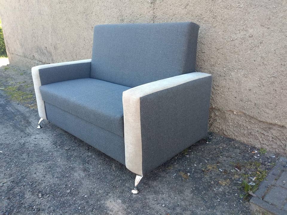 Szara sofa z beżowymi bokami i metalowymi nóżkami, ustawiona na zewnątrz przy szarej ścianie. Widok z boku, lekko z góry. Minimalistyczny design.