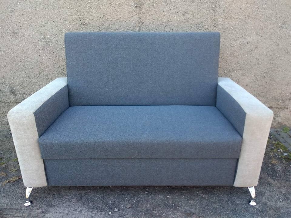 Szara sofa dwuosobowa z jasnymi bokami i metalowymi nóżkami na tle szarej ściany. Prosty, nowoczesny design mebla tapicerowanego.