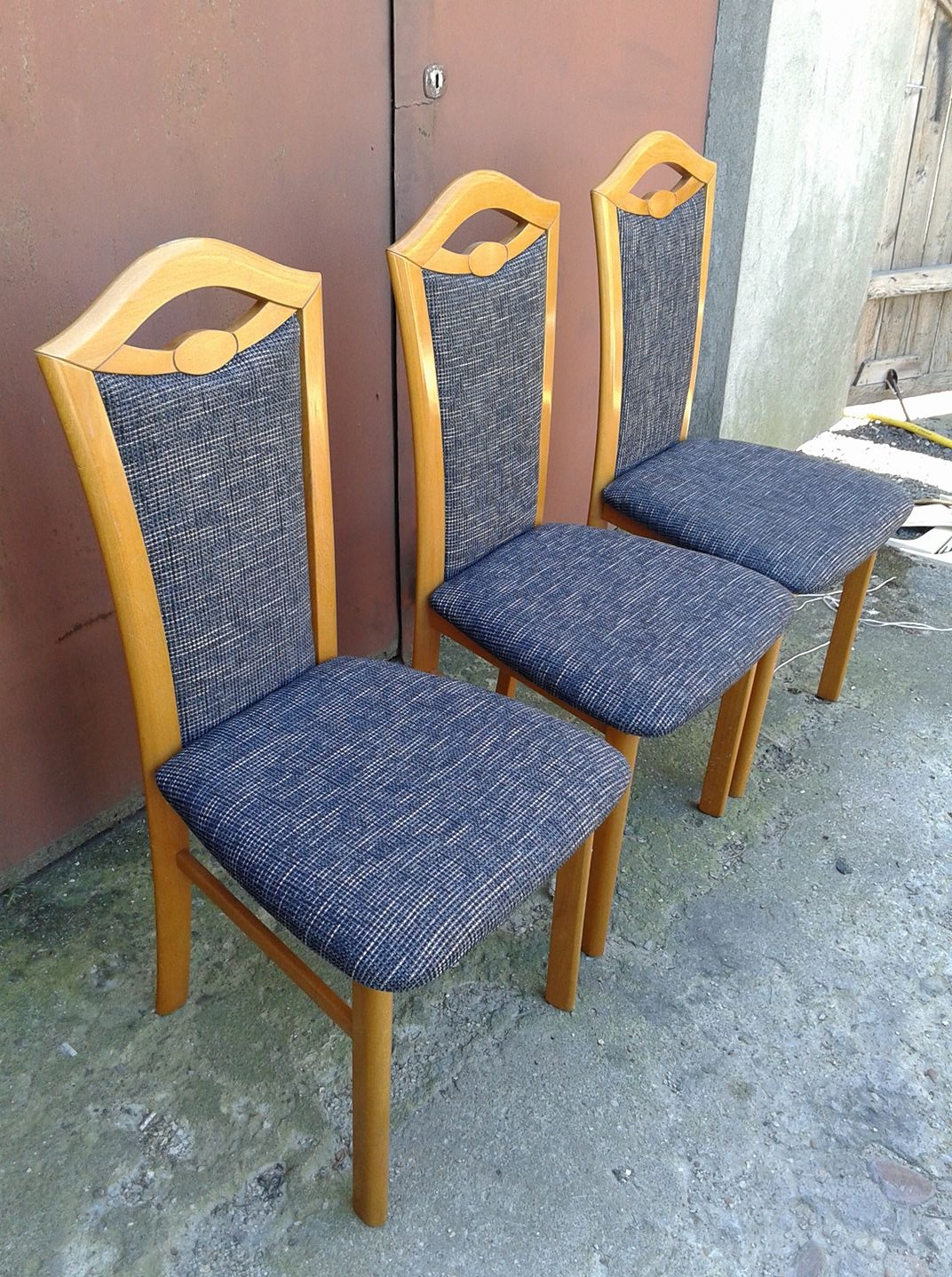 Trzy drewniane krzesła z tapicerowanymi siedziskami i oparciami w odcieniach szarości i błękitu, ustawione na zewnątrz, z widoczną fakturą tapicerki.