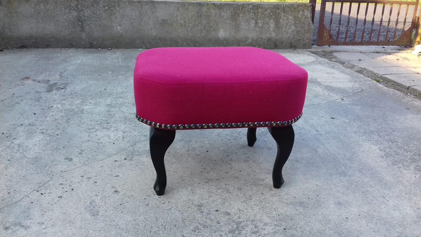 Pufa tapicerowana w odcieniach fuksji, z czarnymi, giętymi nóżkami i ozdobnymi ćwiekami, na tle betonowego podłoża i metalowej bramy.