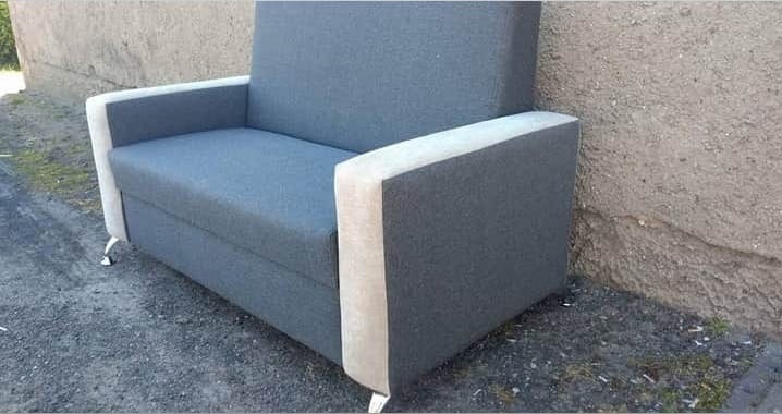 Szary tapicerowany fotel z beżowymi podłokietnikami na metalowych nóżkach, ustawiony na zewnątrz przy szarej ścianie. Widok z boku, lekko z góry.