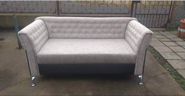 Dwuosobowa sofa w odcieniach szarości z pikowanym oparciem i chromowanymi nóżkami, ustawiona na zewnątrz na betonowej nawierzchni. Widoczne ogrodzenie w tle.